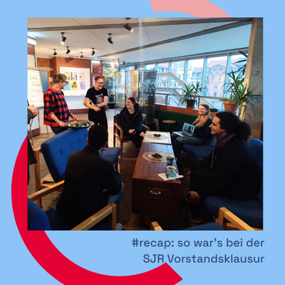 eine Gruppe von Menschen sitzt auf einer Sitzmöbelgruppe um einen niedrigen Tisch. Links steht eine Frau mit einem Tablett, auf dem sich verschiedene Gegenstände befinden. rechts neben ihr steht eine weitere Person. Im Hintergrund erkennt man ein Flipchart und eine Fensterfront.